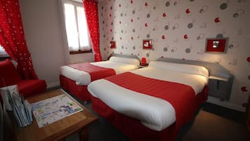 Habitación de hotel con dos camas, una mesa de madera con revistas, cortinas rojas y una pared con diseño circular.