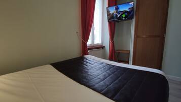 A hotel room with a bed, a television mounted on the wall, a window with red curtains, and a radiator.