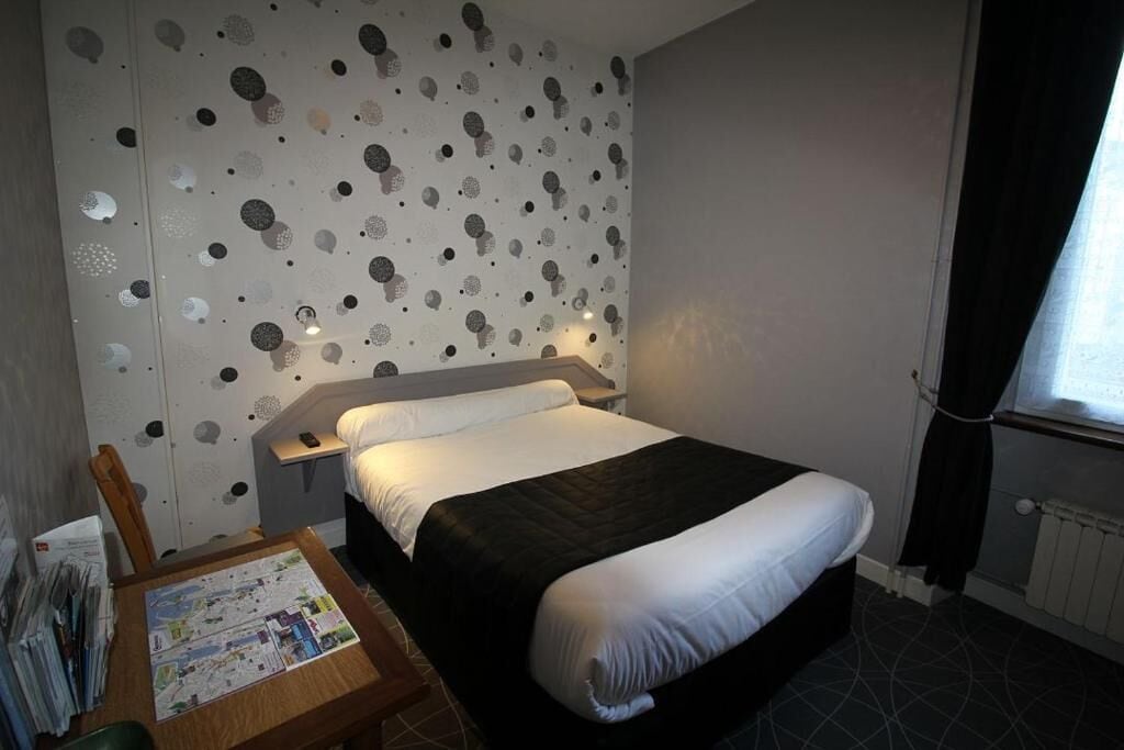 Habitación de hotel con cama, escritorio con papeles, pared con estampado de lunares y ventana con cortinas.