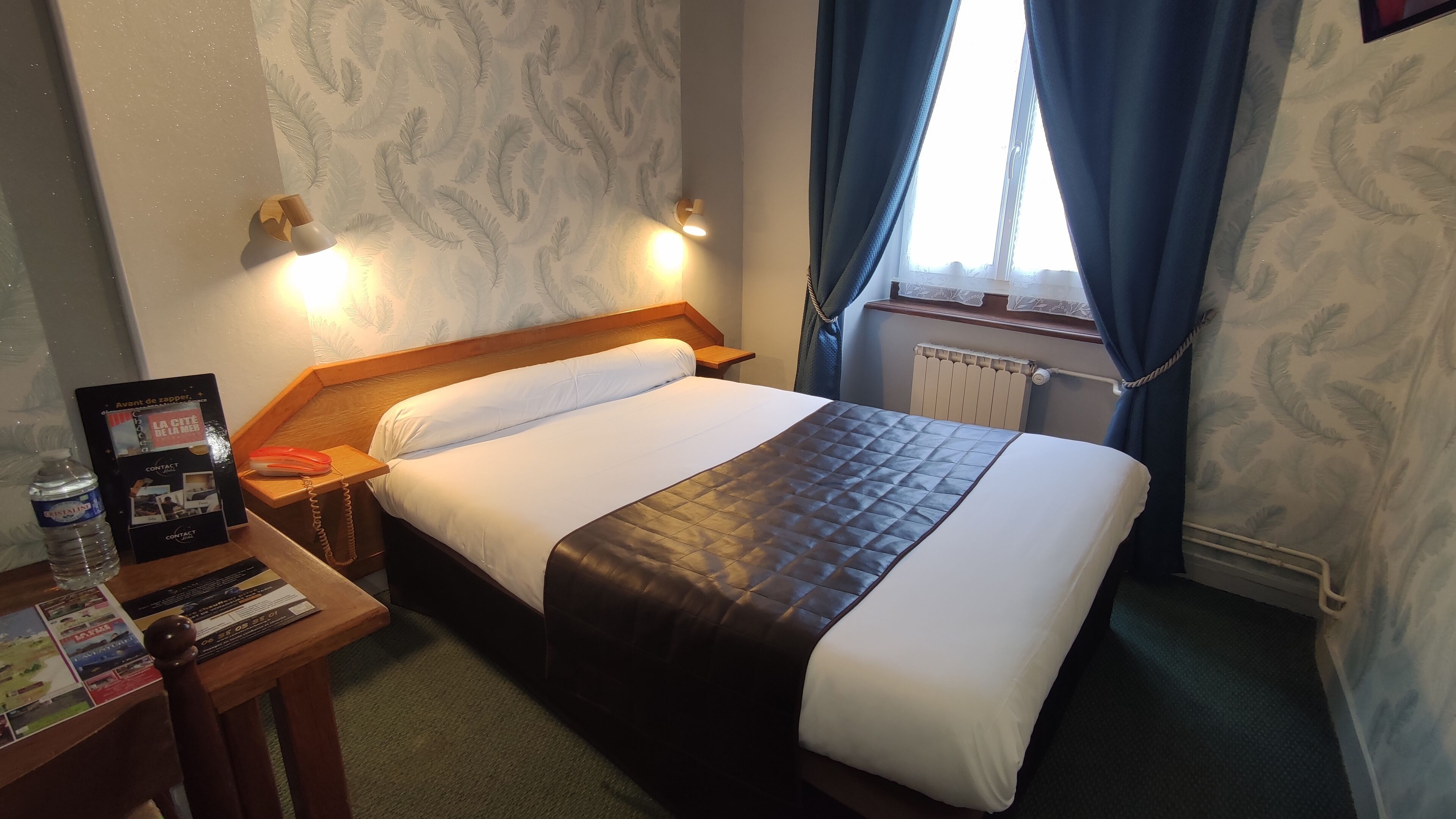 Een hotelkamer met een bed, nachtkastje met lamp, telefoon en een brochure.