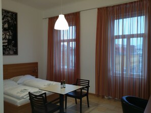 A hotel room with a bed, a table with chairs, a hanging light fixture, and large windows with curtains. - Hotel Aton (Graz)