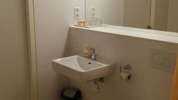 A small bathroom with a white sink, a toilet, and a mirror above the sink. There is a trash can next to the sink and a light above the mirror.