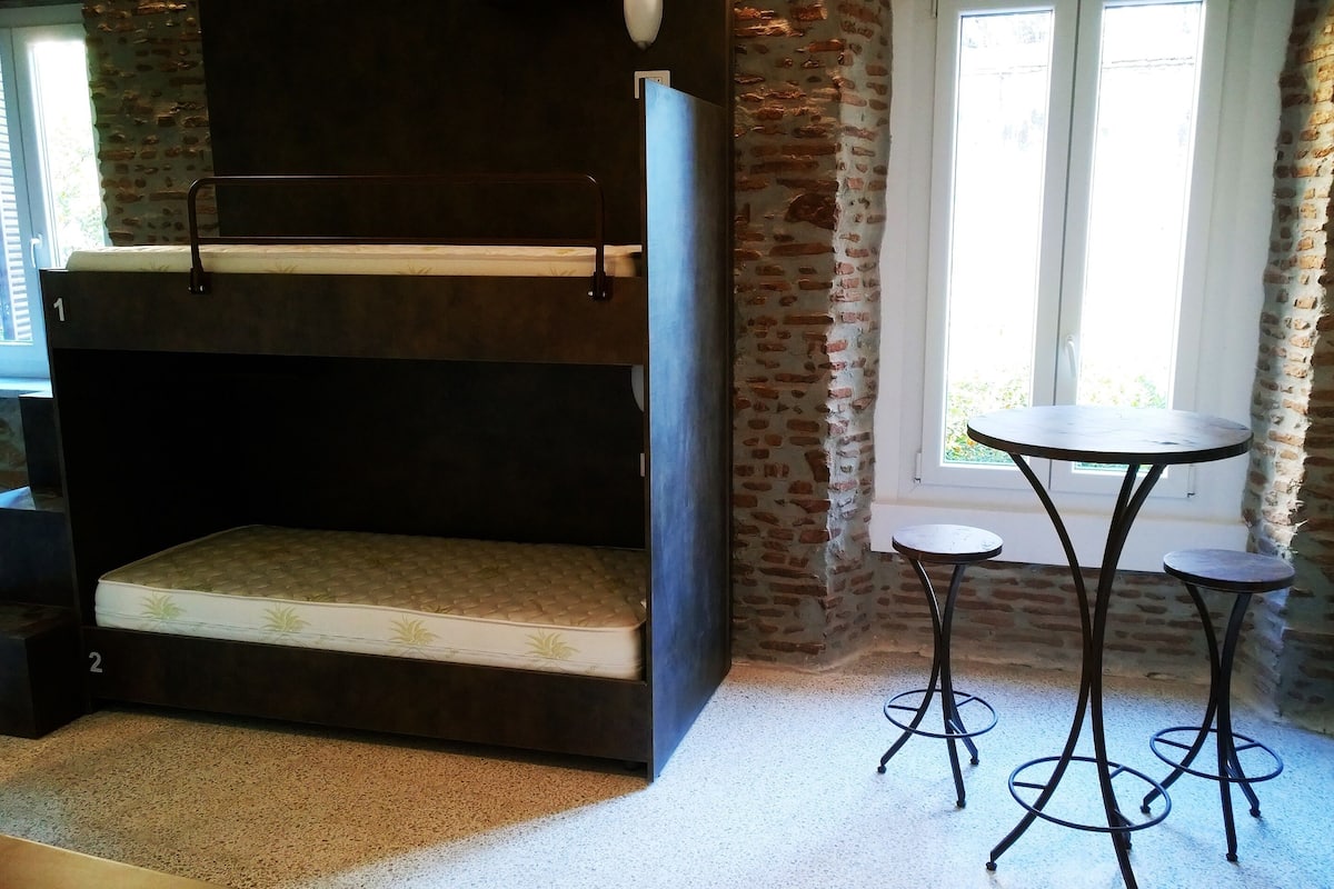 A bunk bed with a mattress, a small table with stools, and a window with curtains.