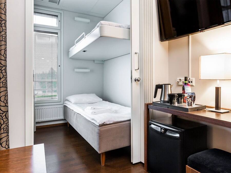Ein kompaktes Hotelzimmer mit Etagenbett, kleinem Schreibtisch mit Lampe und einem Fenster mit Jalousien.