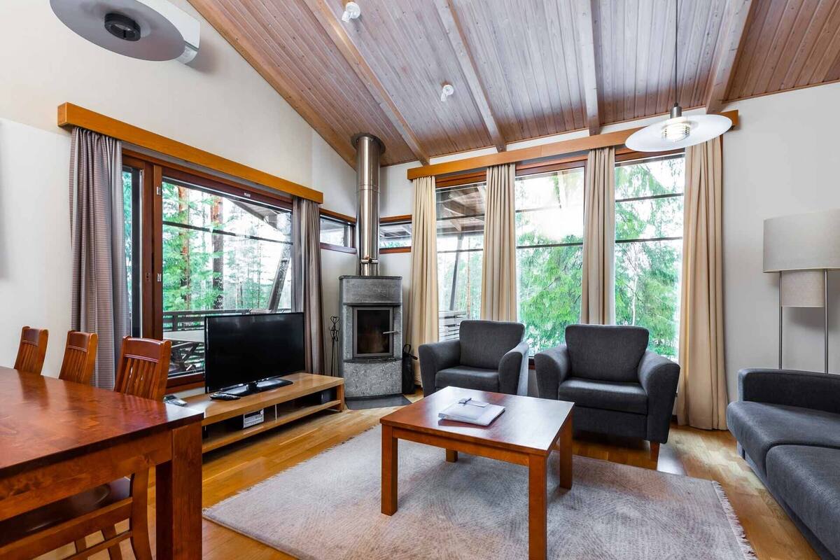 A cozy living room with a fireplace, wooden furniture, and large windows offering a view of the forest.