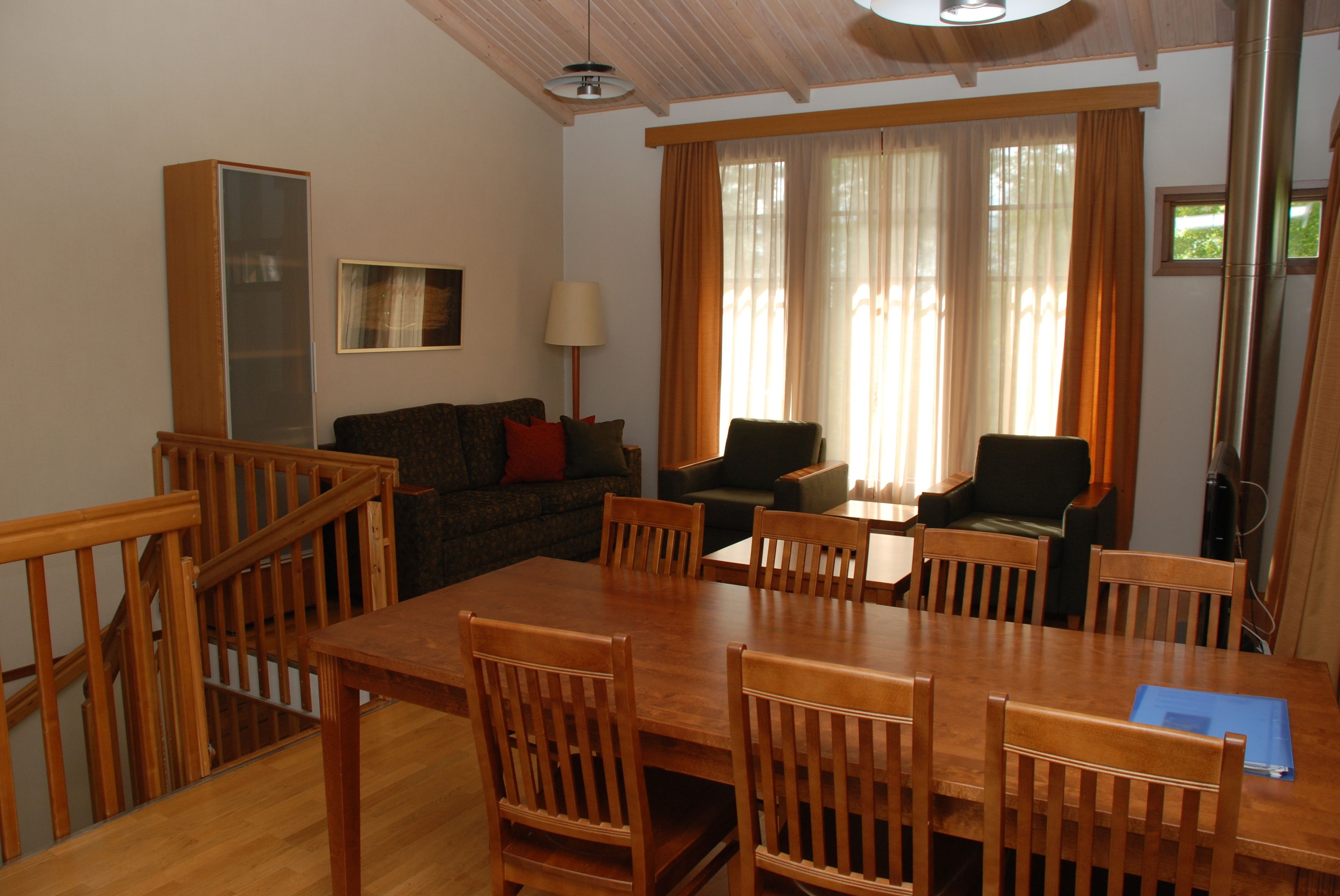 Un comedor con mesa y sillas de madera, un sofá con cojines, una lámpara y un amplio ventanal con cortinas.