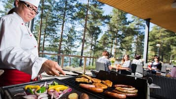 A chef grilling sausages and vegetables outdoors with a forest view.