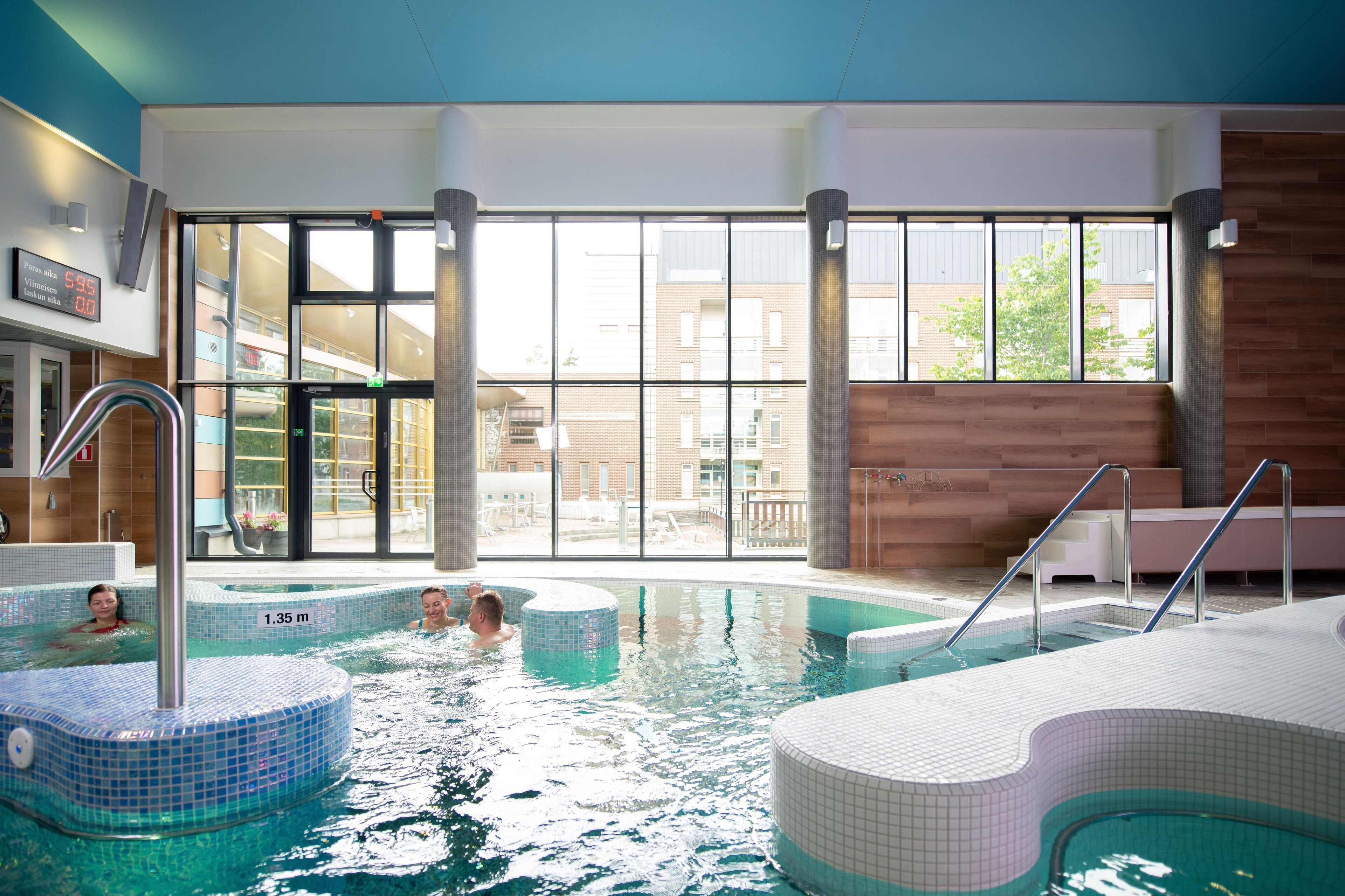 Una piscina interior con agua cristalina, un indicador de profundidad visible y vistas de los edificios exteriores a través de amplios ventanales.