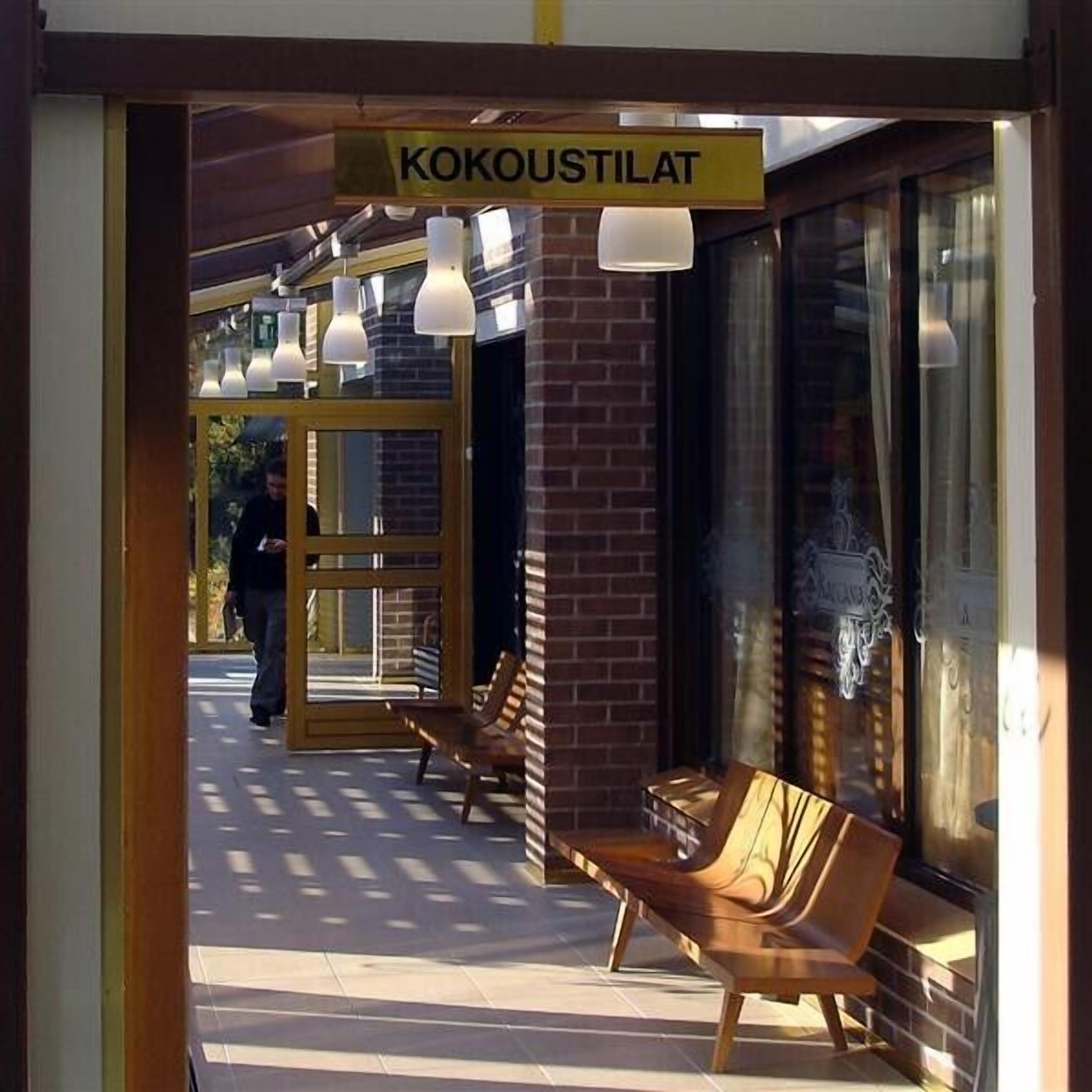 Entrada de um café com uma placa indicando "KOKUSTILAT". Há bancos de madeira do lado de fora, uma porta de vidro e luminárias penduradas.