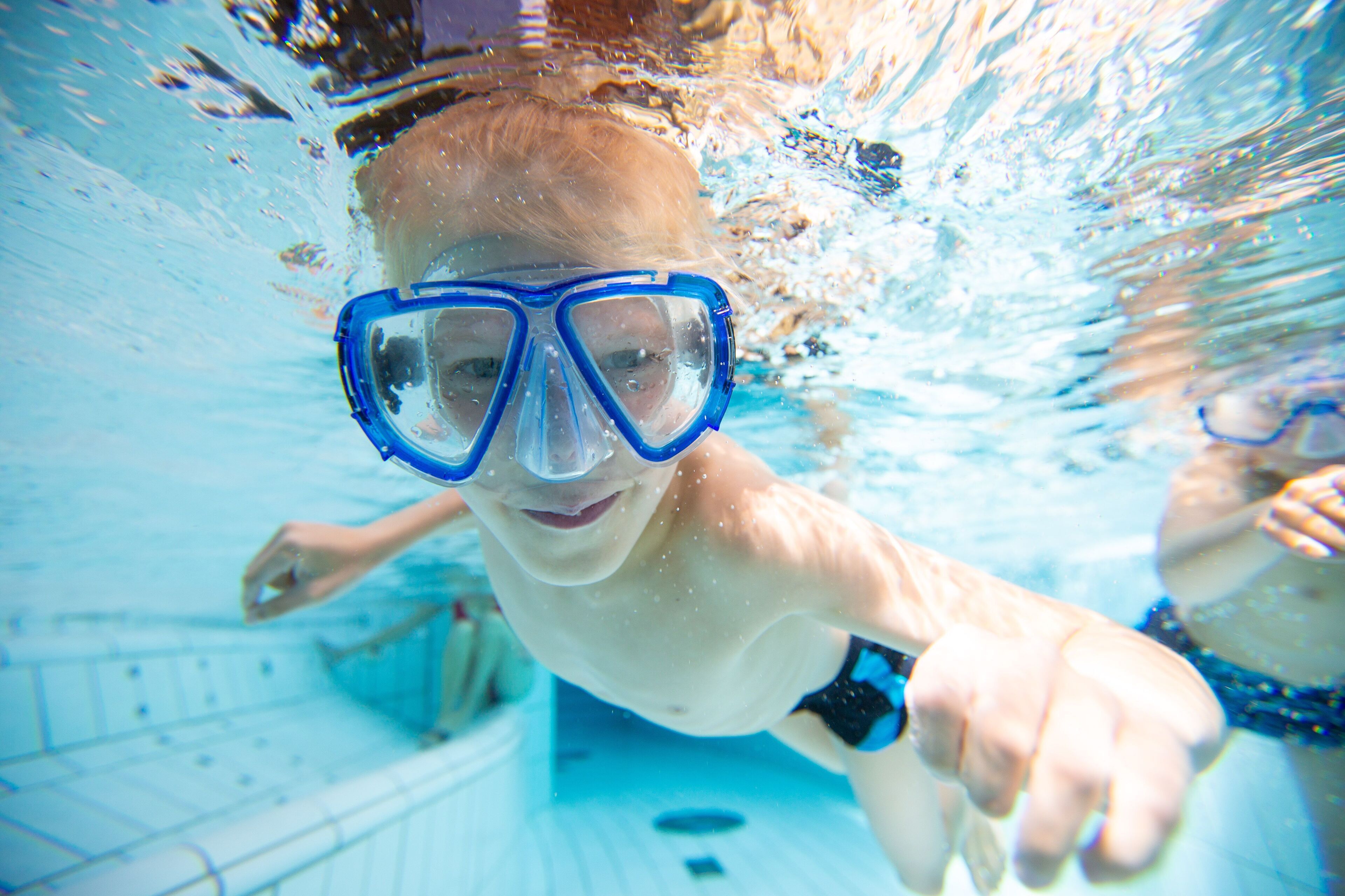 Une vue sous-marine d'une piscine avec de l'eau claire, une voie de nage bien visible et un nageur portant des lunettes de natation bleues.