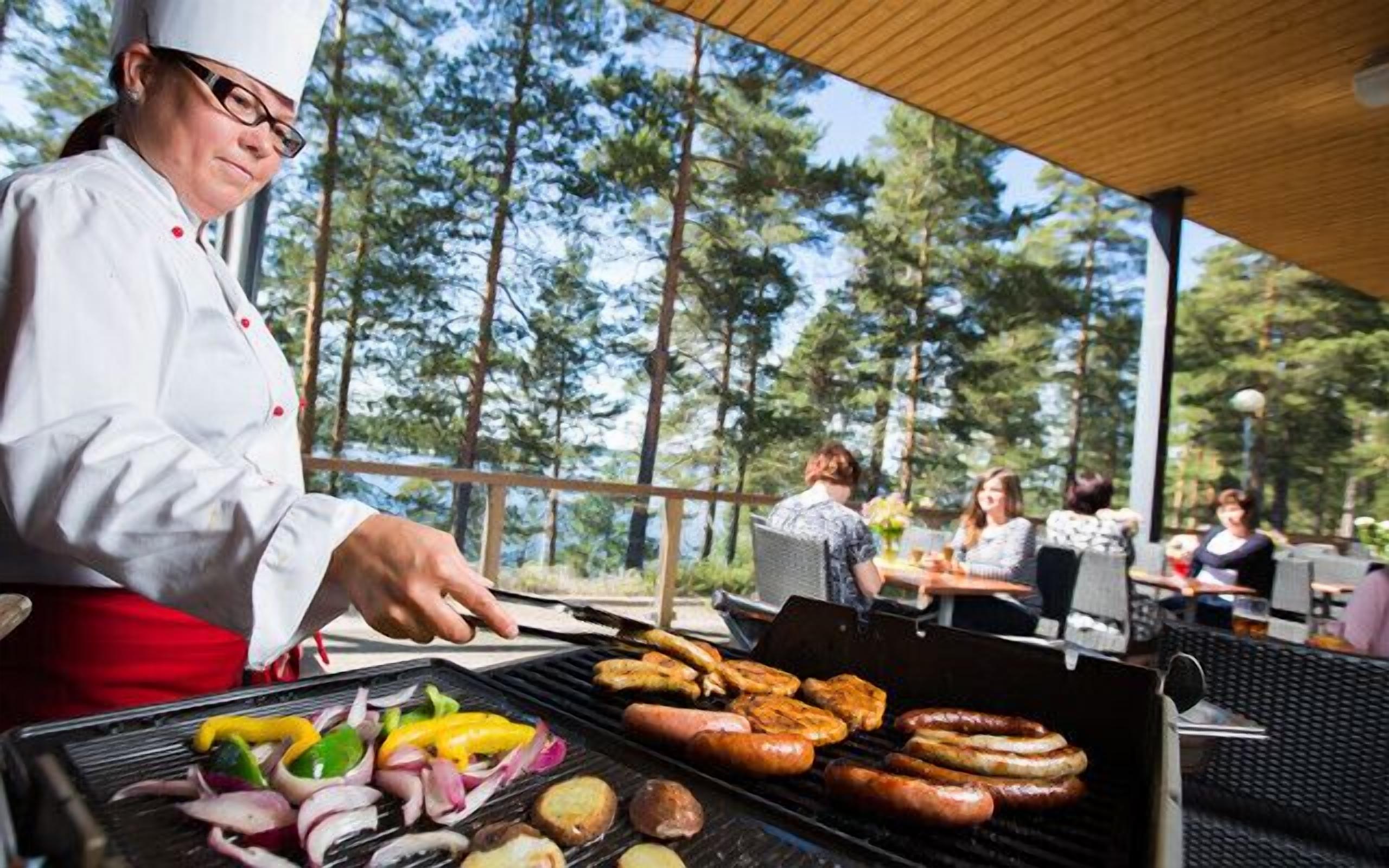 Un cocinero asando salchichas y verduras al aire libre, con vistas a un bosque.