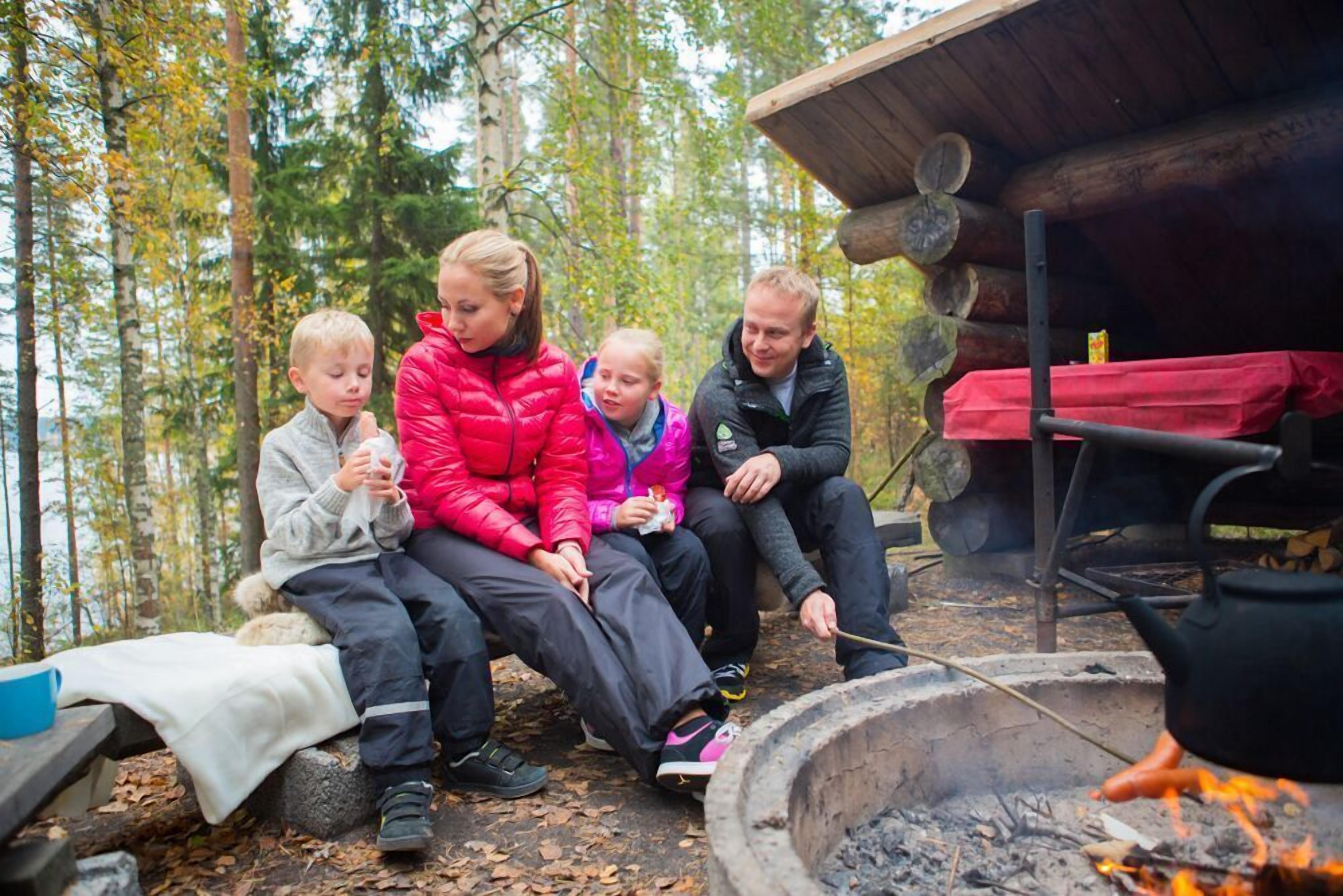 Ryhmä ihmisiä istuu nuotion ympärillä metsäisellä alueella, ja taustalla näkyy puinen mökki.