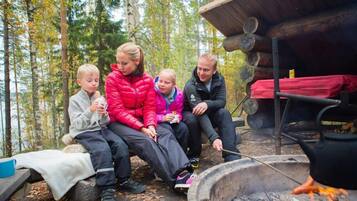 Ryhmä ihmisiä istuu nuotion ympärillä metsäisellä alueella, ja taustalla näkyy puinen mökki.