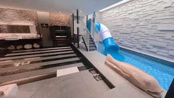 A modern indoor pool area with a blue slide, a stone wall, and a staircase leading to a higher level.