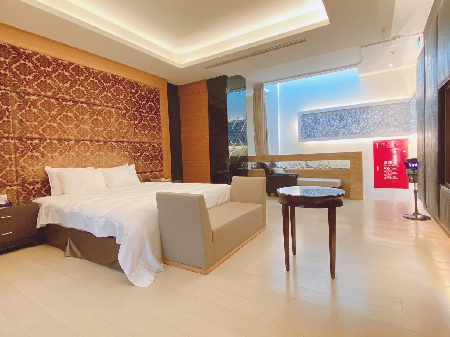 A modern hotel room with a large bed, a sofa, a small table, and a view of the sea through a large window.