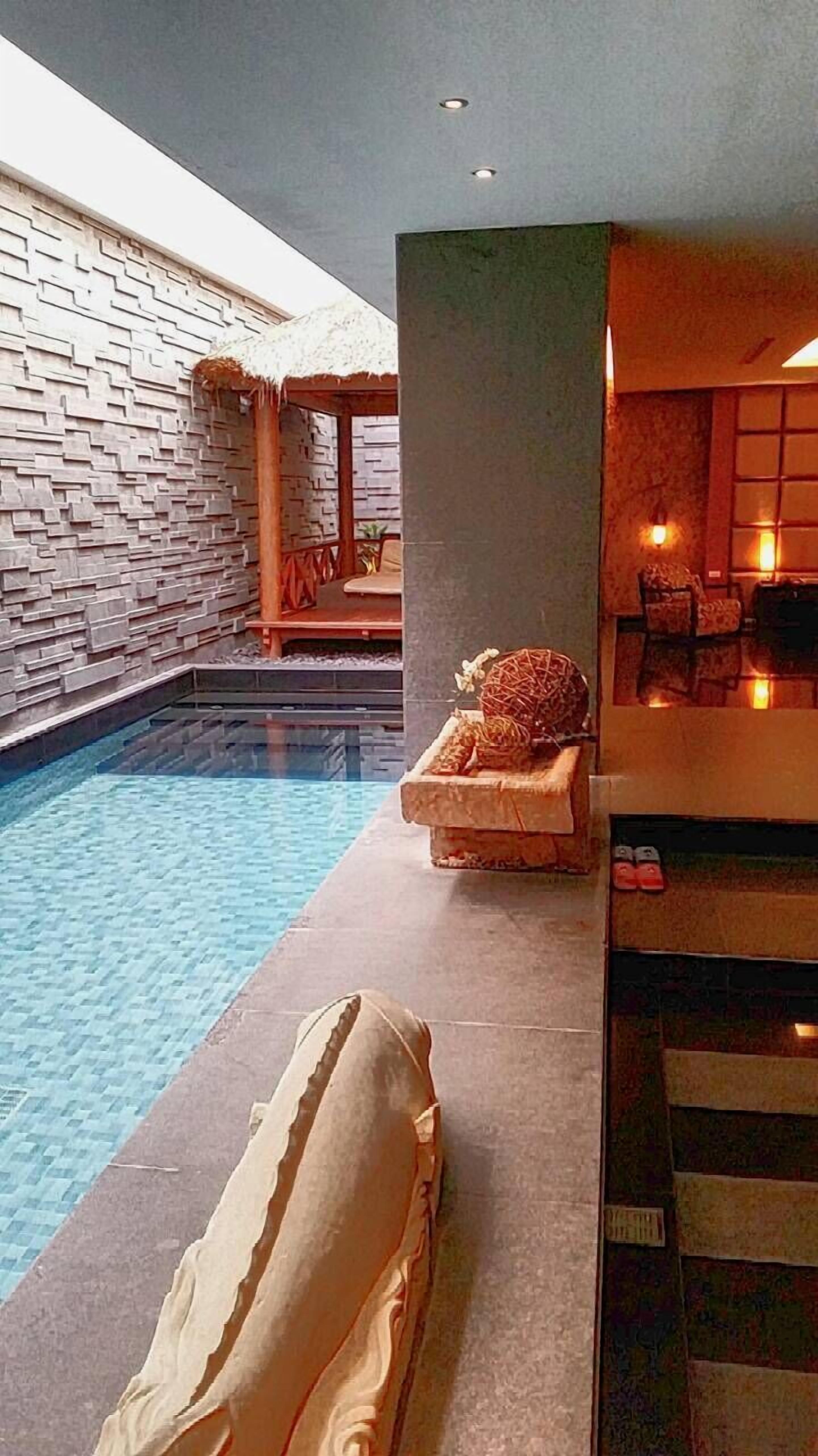 A modern indoor pool area with a stone wall, a lounge chair, and a small seating area with cushions and a table.