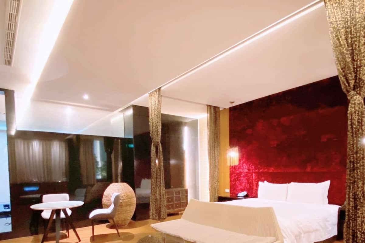 Quarto de hotel moderno com cama, sofá, mesinha e cadeira. Possui uma janela grande e uma parede com detalhes em vermelho.