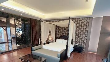A four-poster bed with a canopy, a small table, and a sofa in a modern hotel room with patterned wallpaper and a large window.