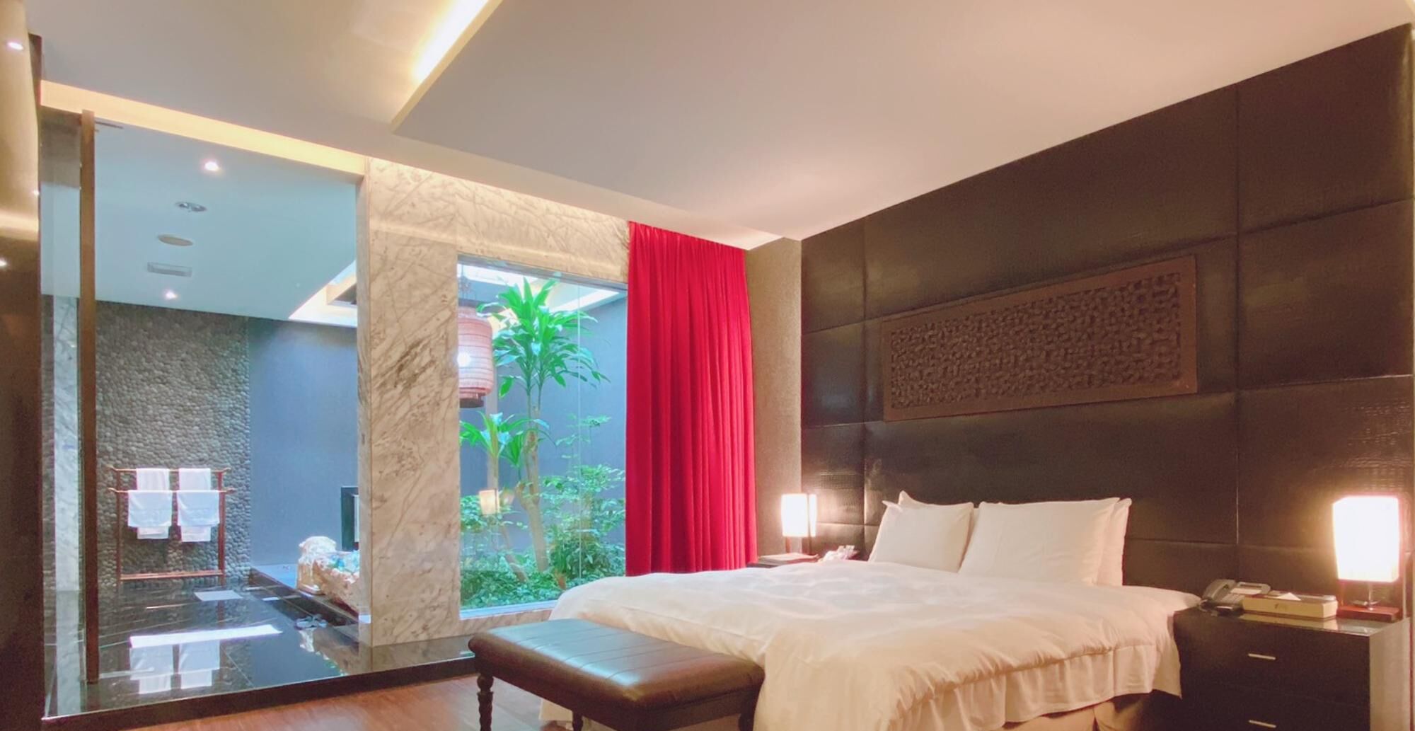 Habitación de hotel moderna con una cama grande, una mesita de noche con lámpara, un espejo y vistas a un jardín a través de una puerta de cristal.