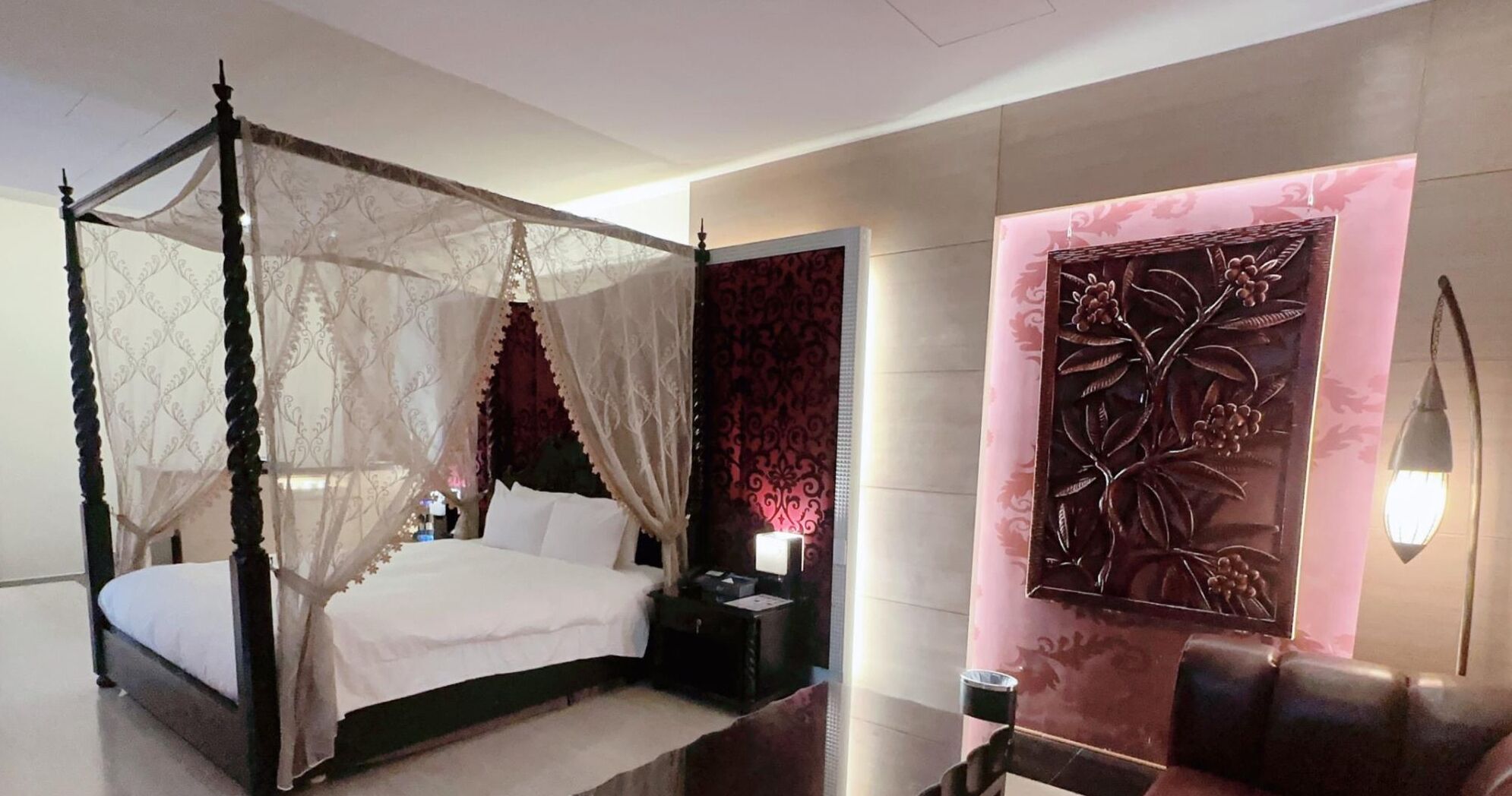 A four-poster canopy bed with a nightstand beside it, a wall-mounted lamp, and a decorative panel with floral carvings.