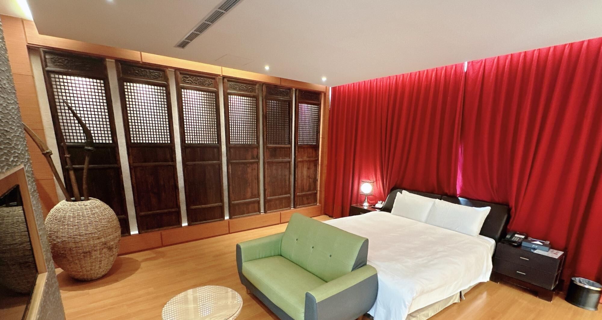 A modern hotel room with a large bed, a green armchair, a wooden floor, and a traditional-style window with red curtains.
