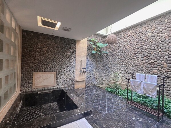 Un área de spa con pared de piedra, suelo de mármol negro y un pequeño jardín con plantas, incluyendo una planta colgante.