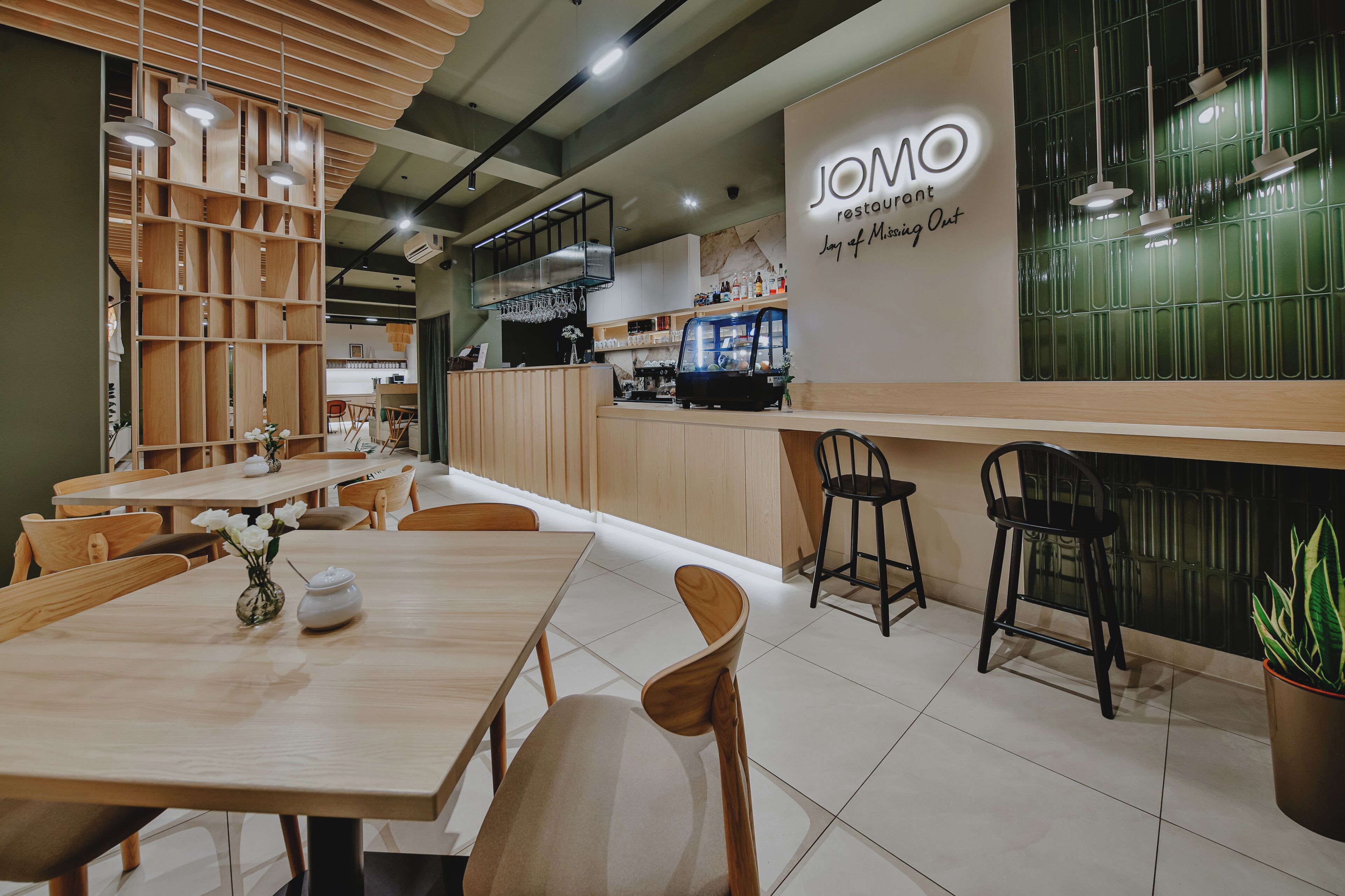 A restaurant interior with wooden tables, black bar stools, a counter, and a wall sign reading 'JOMO restaurant.'