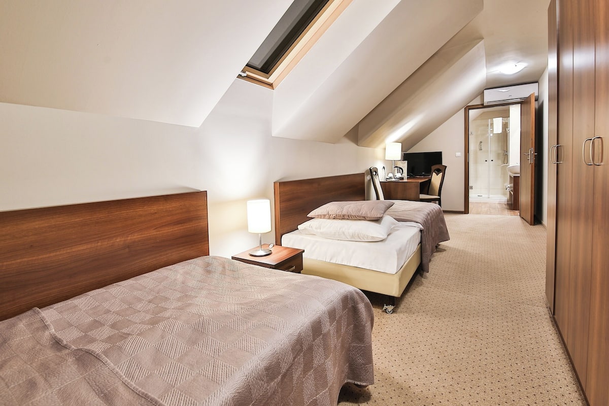 A hotel room with a sloped ceiling, two beds, a desk, a chair, a lamp, and a wardrobe.