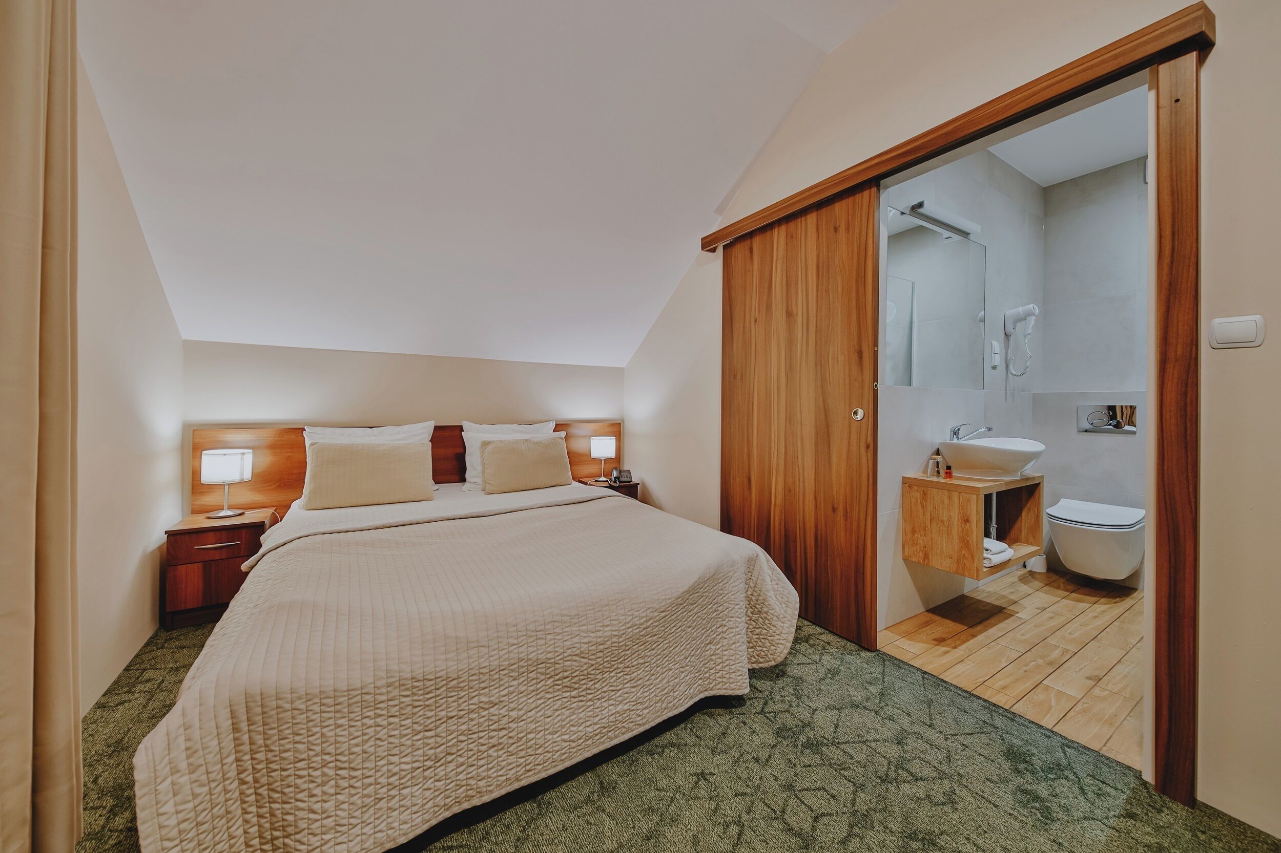 Habitación de hotel con cama, mesitas de noche, armario de madera y baño con lavamanos e inodoro.