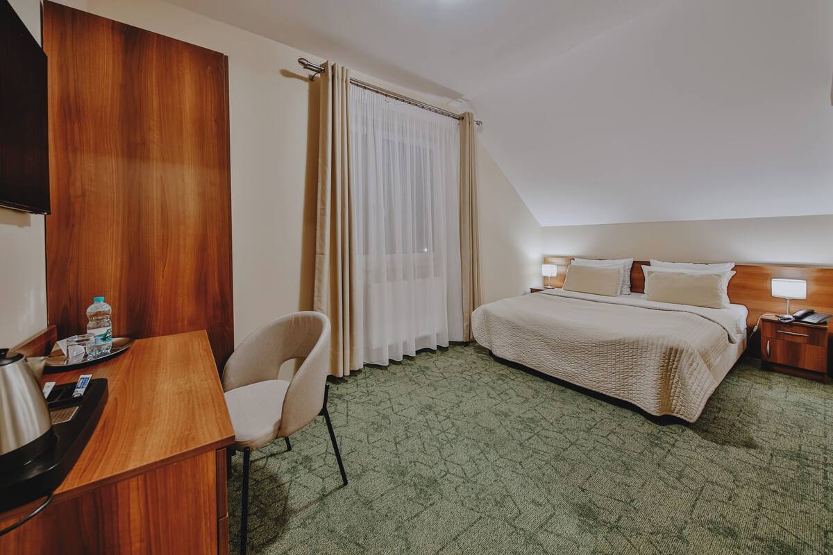 A hotel room with a bed, a desk with a chair, a TV, a bedside table with a lamp, and a window with curtains.