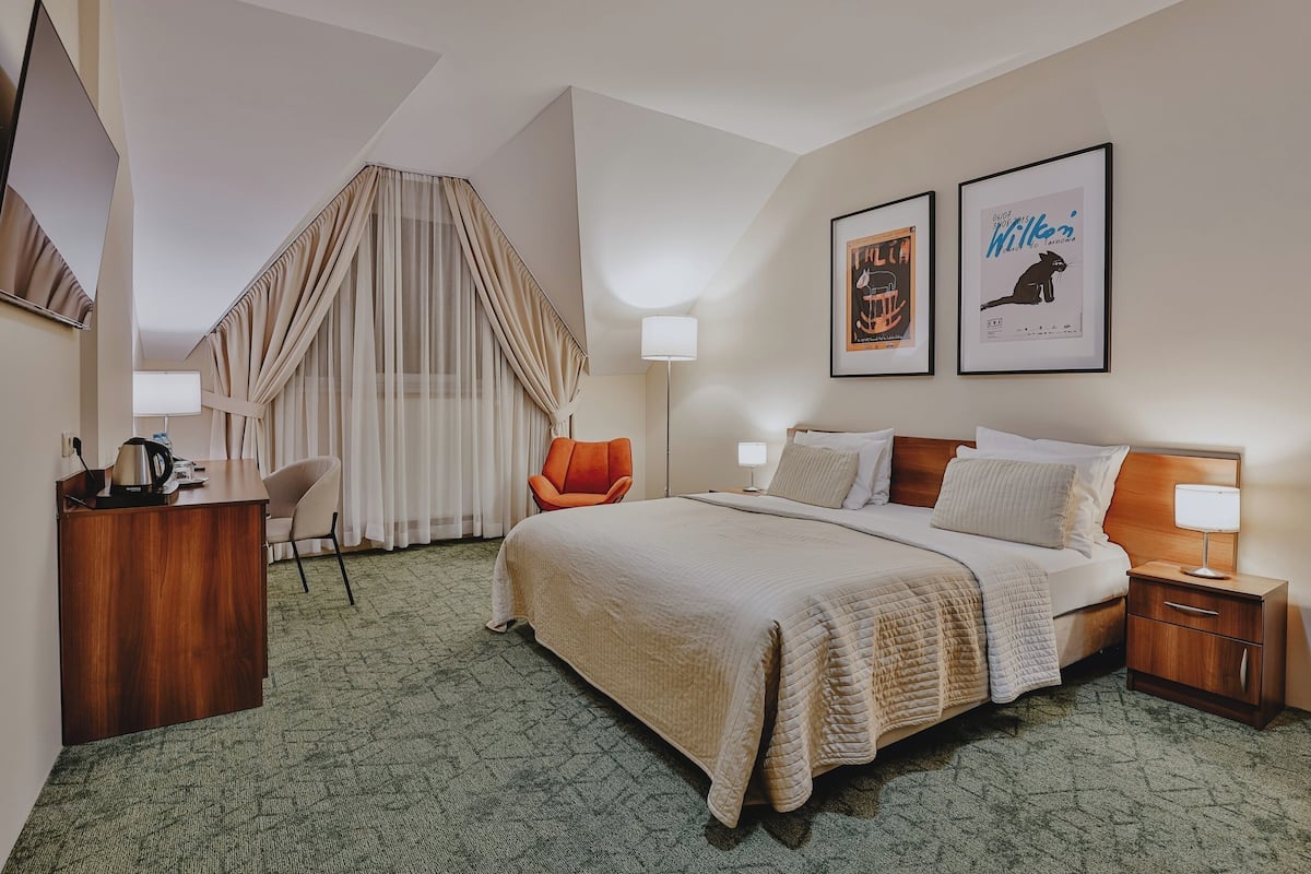 A hotel room with a bed, desk, chair, and two framed pictures on the wall.
