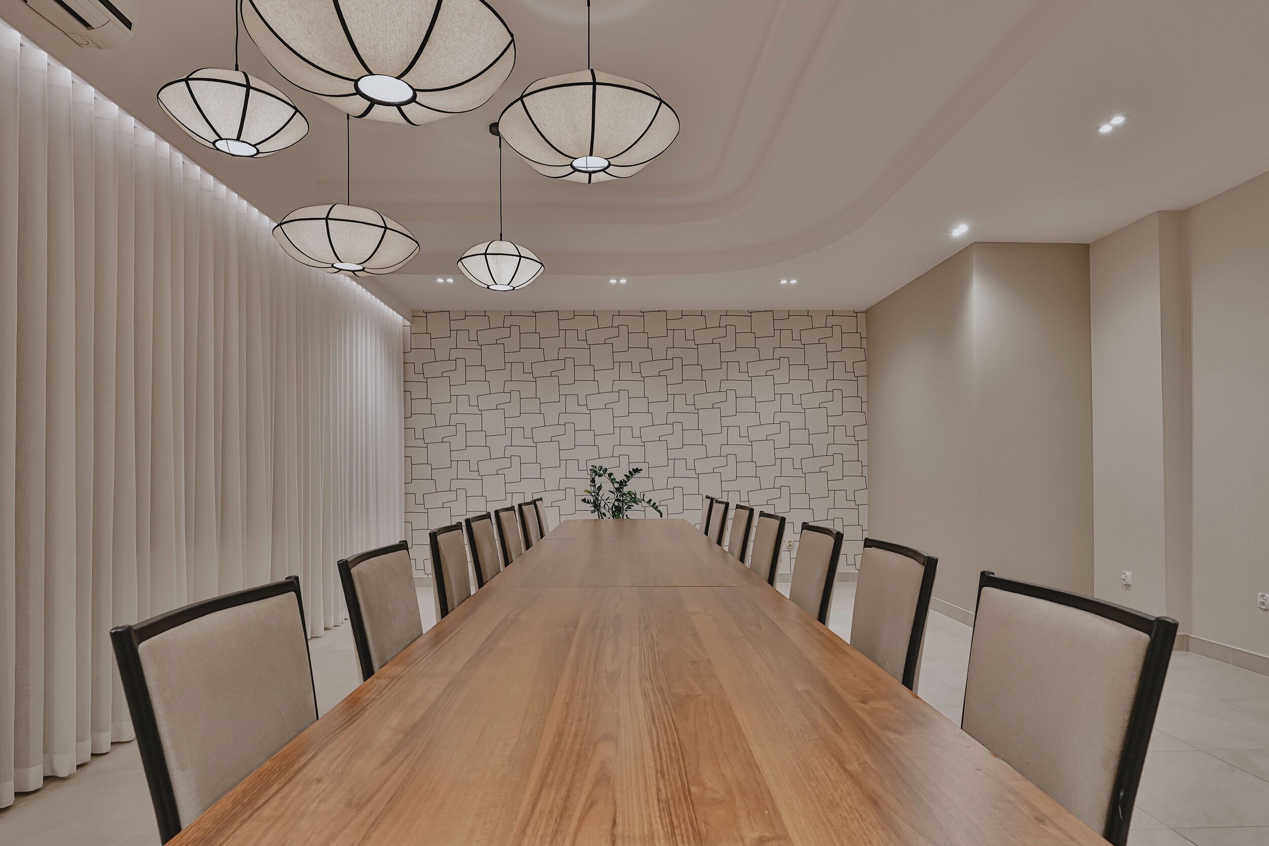 A conference room with a long wooden table, multiple chairs, hanging pendant lights, and a patterned wall.