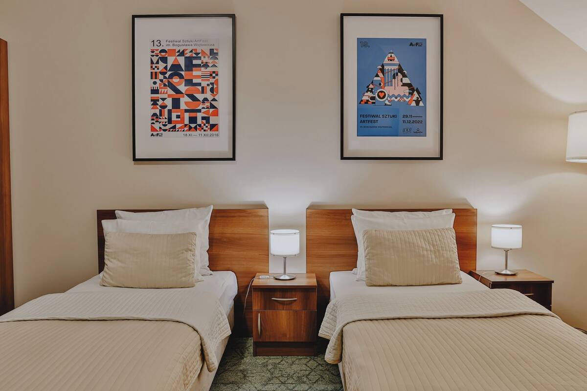 A hotel room with two beds, wooden headboards, bedside tables with lamps, and framed posters on the wall.