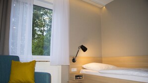 A hotel room with a bed, a bedside table, a lamp, a window with curtains, and a sofa with a pillow. - Haus Mariengrund (Münster)