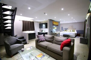 A modern hotel room with a sofa, armchair, glass coffee table, and a bed with a headboard.