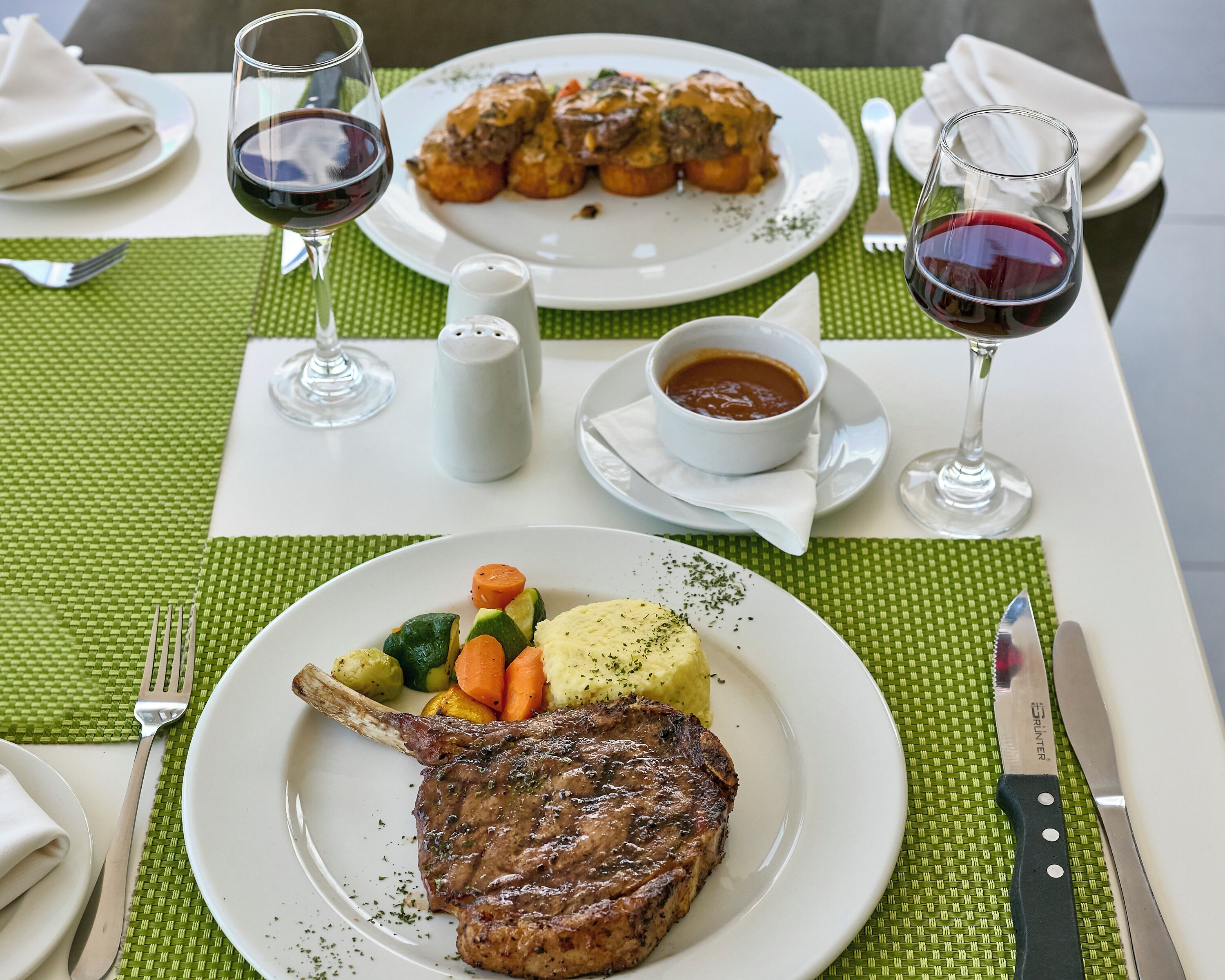 A dining table with a steak, vegetables, and a bowl of sauce. There are two glasses of red wine and a salt shaker on the table.
