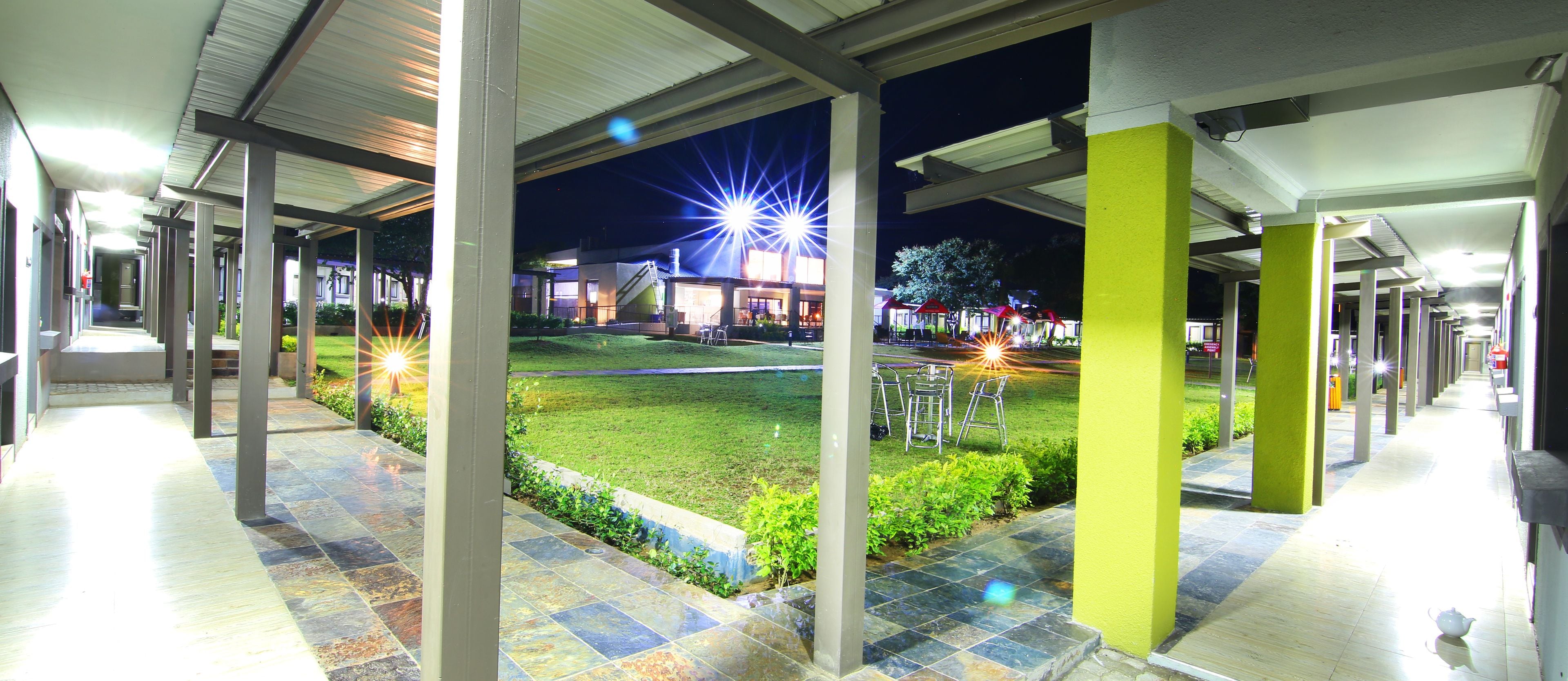 A covered outdoor area with a tiled floor, green landscaping, and bright lights in the background.