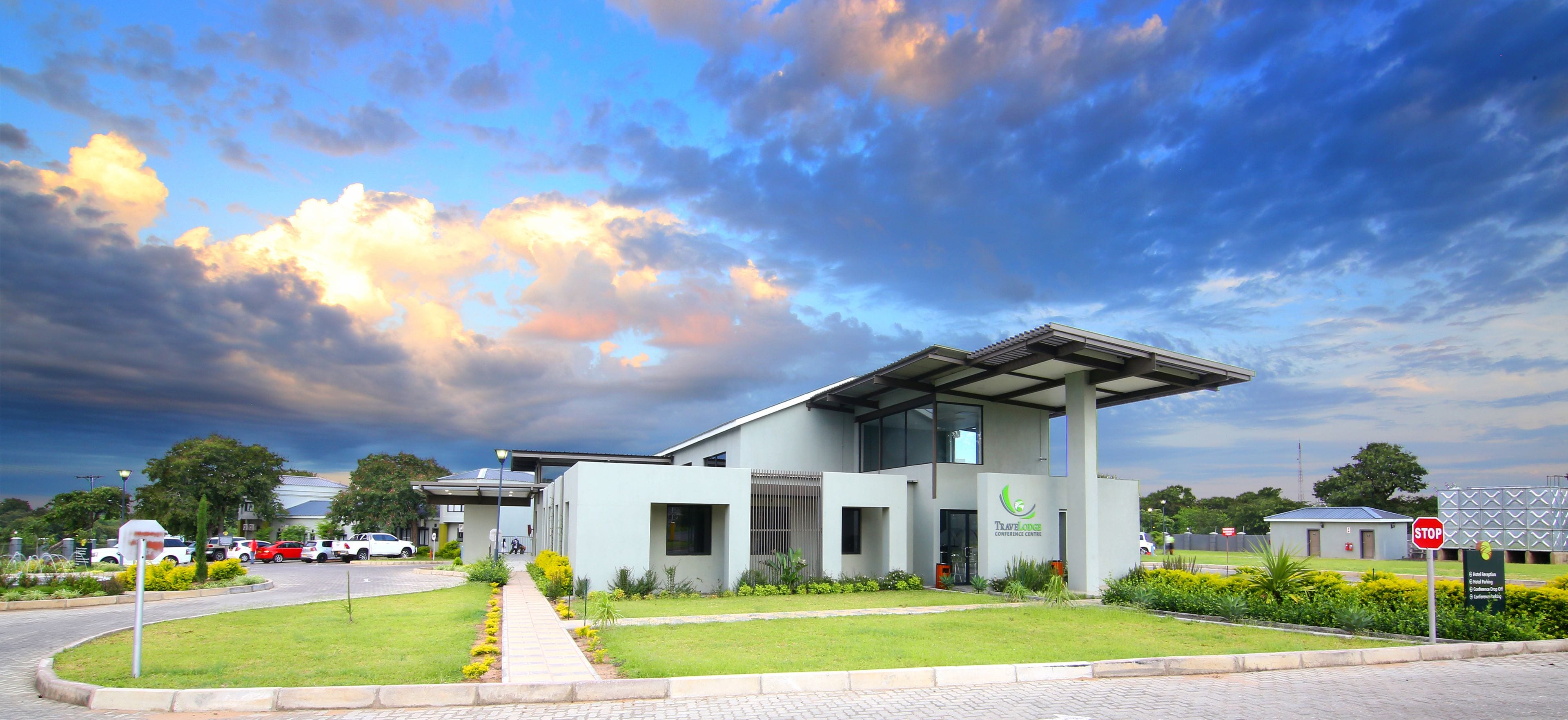 A modern building with a flat roof and large windows, surrounded by a well-maintained lawn and a parking area with several cars.