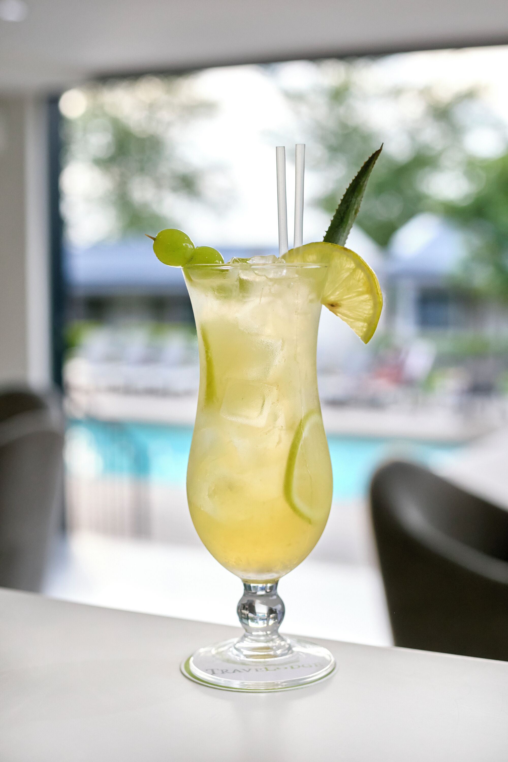 A tall glass with a lime slice and a mint leaf garnish, placed on a table with a view of a pool and outdoor seating.