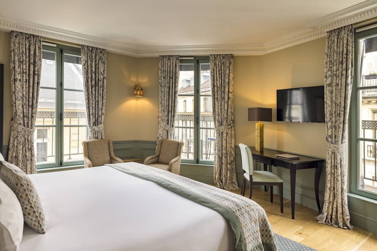 Habitación de hotel con una cama grande, dos sillones, un escritorio con silla, un televisor de pantalla plana y una ventana con cortinas.