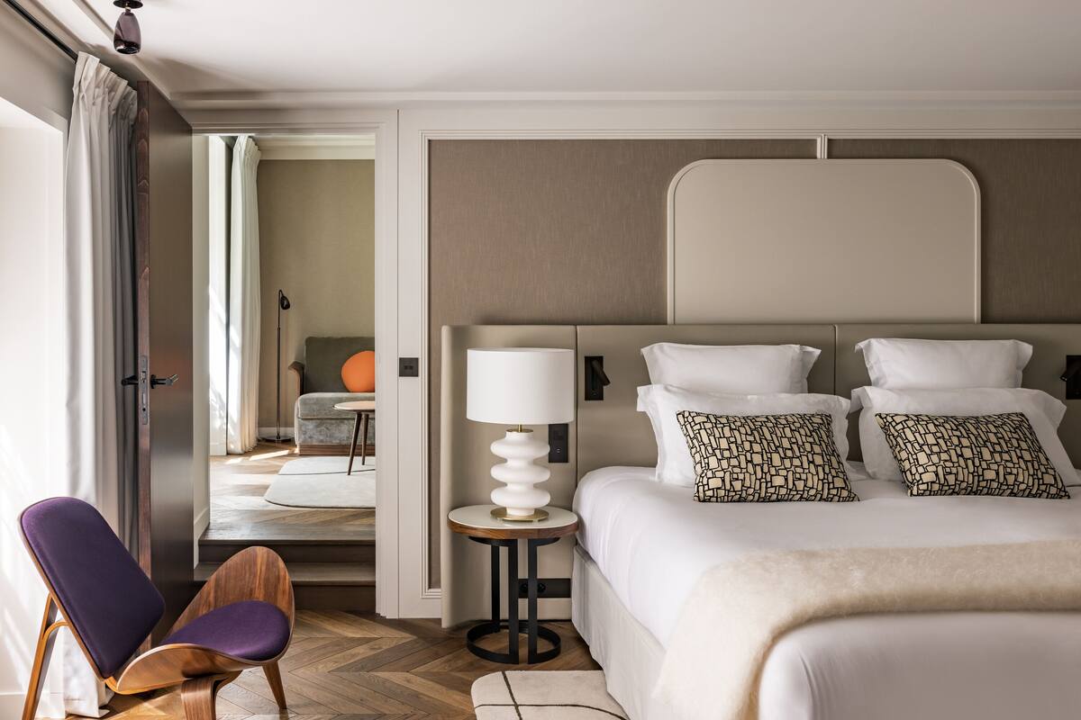 Habitación de hotel moderna con una cama grande, una mesita de noche con lámpara, una silla y vista a un comedor.