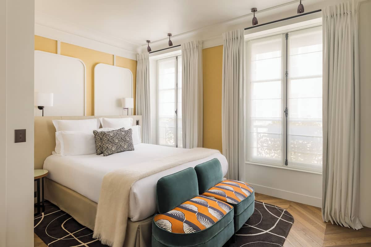 Una habitación de hotel con cama, un banco, una ventana con cortinas y una lámpara.