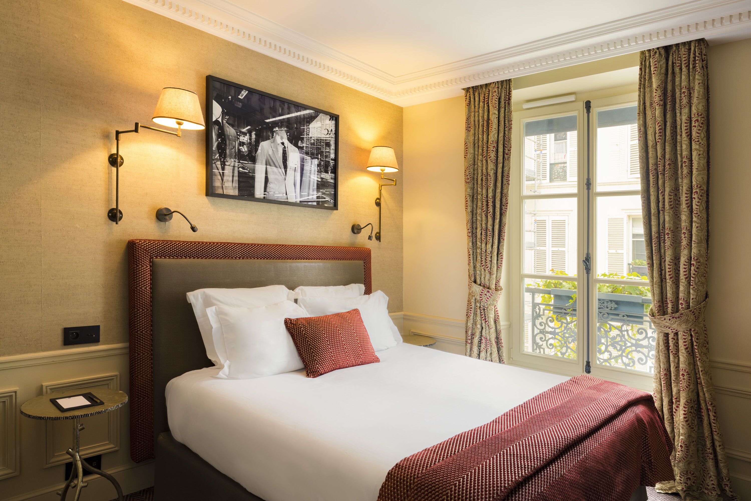 Habitación de hotel con una cama grande, dos lámparas de pared, un cuadro enmarcado sobre la cama y una ventana con cortinas.