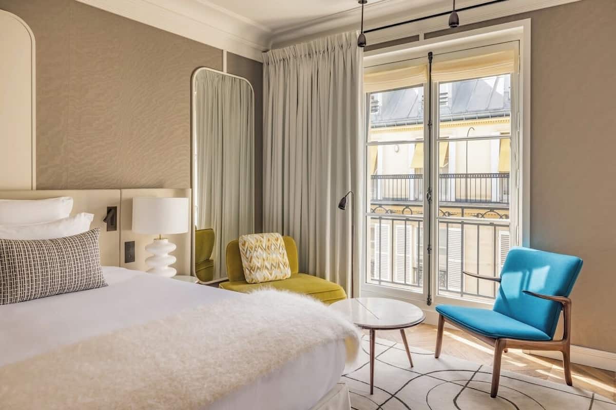 Habitación de hotel con una cama grande, una mesita redonda pequeña, un sillón azul y un balcón con vistas a la ciudad.