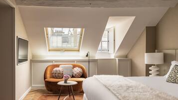 Quarto moderno com uma cama grande, uma mesa redonda pequena com um vaso, uma poltrona marrom e uma janela com vista para edifícios.