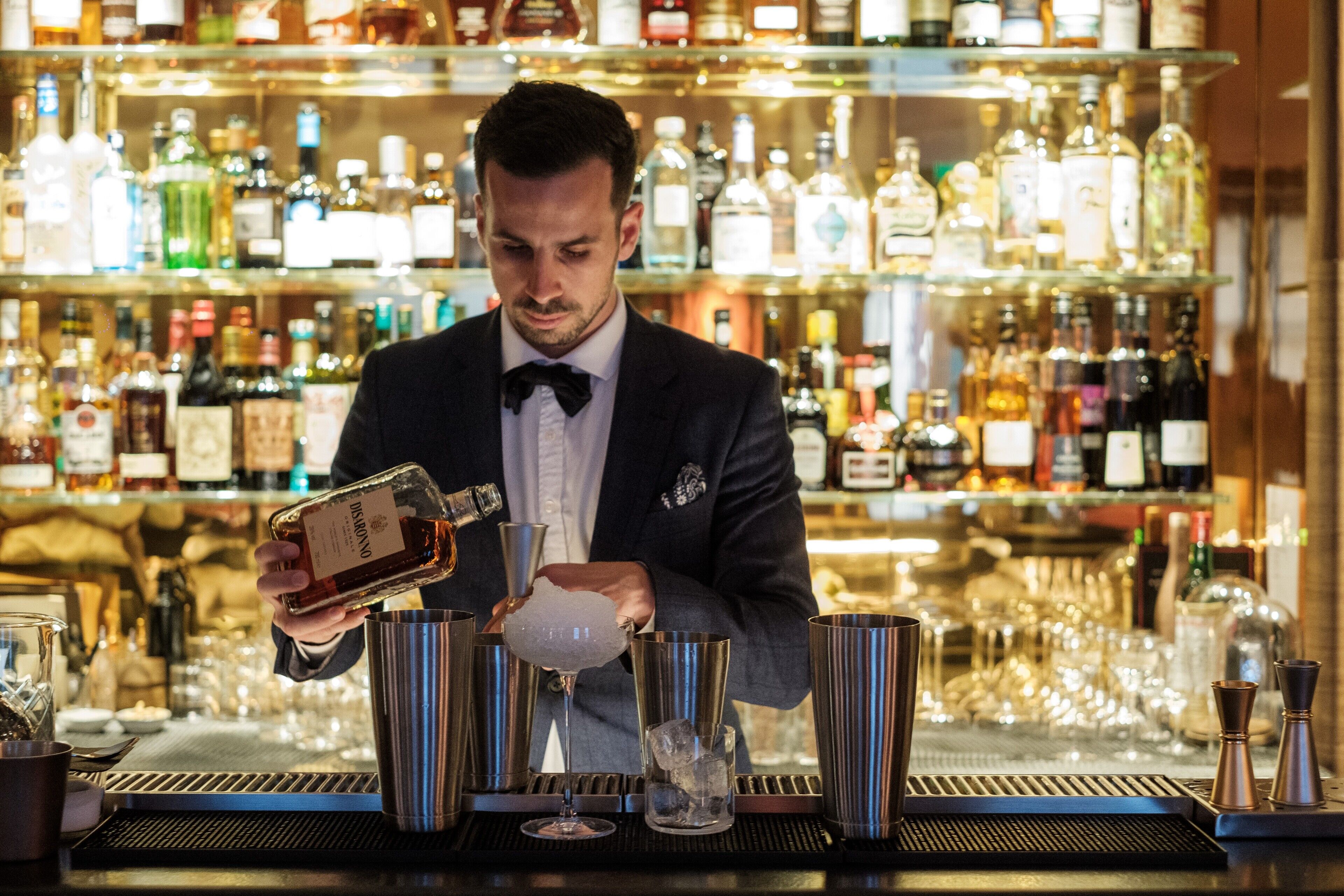 A bartender preparing a drink at a bar with a variety of bottles in the background.