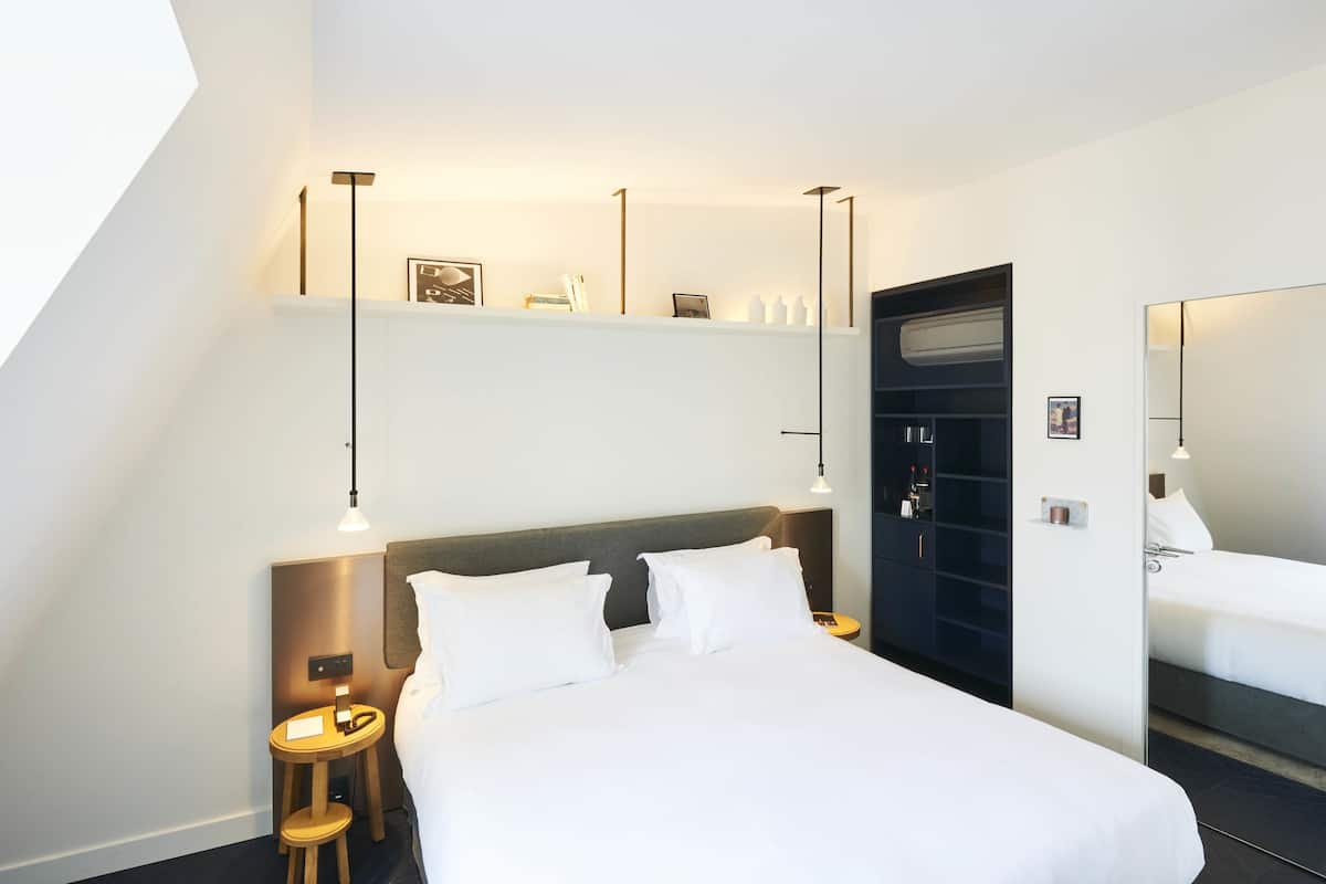 Una habitación de hotel moderna con una cama grande, mesitas de noche, un taburete pequeño, un espejo y una estantería con adornos.