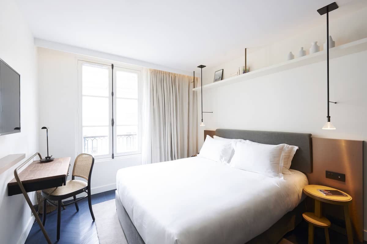 Habitación de hotel con cama, escritorio con silla, ventana con cortinas y mesita de noche con lámpara.