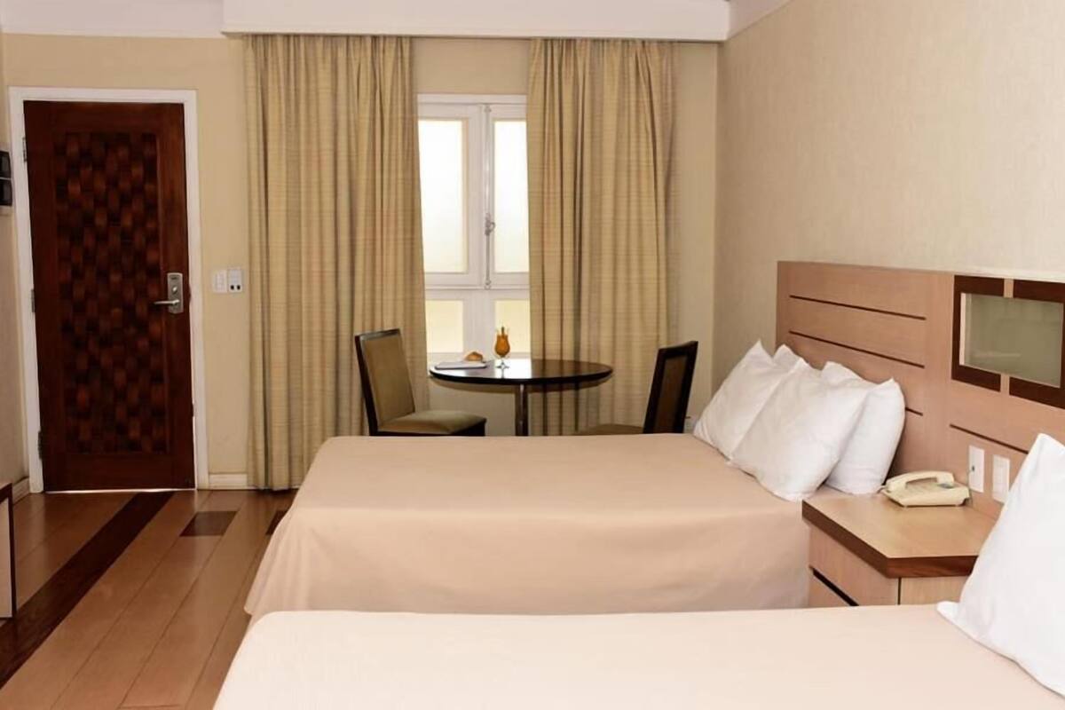 A hotel room with two beds, a small table, chairs, a window with curtains, and a door.