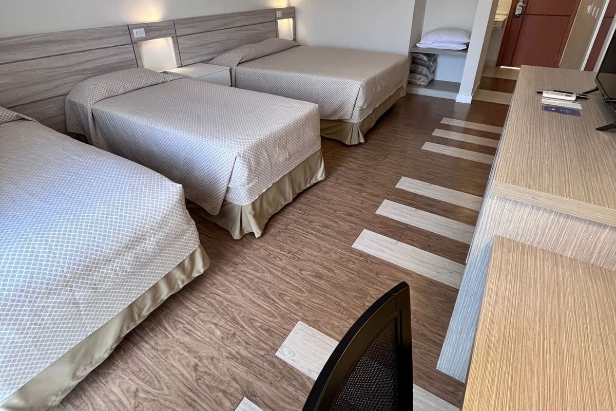 A hotel room with three beds, a desk, and a chair. There is an air conditioning unit on the wall and a door leading to another room.